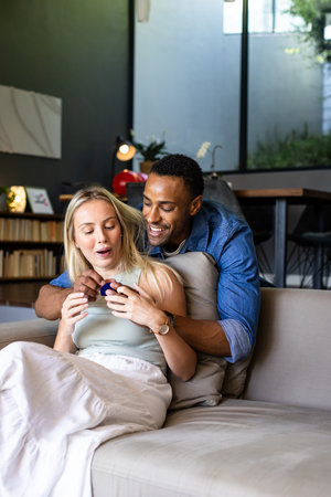 Couple at home, man surprising woman with gift, both smiling and joyful. Romance, celebration, surprise, happiness, relationship, cheerfulの写真素材