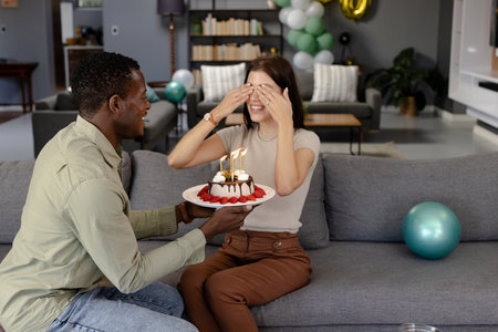 African American man surprising woman with birthday cake, both smiling on sofa. Celebration, surprise, couple, happiness, living room, relationshipの写真素材