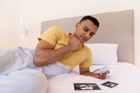 Young man lying on bed, thoughtfully looking at ultrasound photos. Pregnancy, parenthood, expecting, contemplation, fatherhood, futureの写真素材
