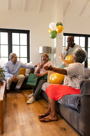 Elderly friends celebrating birthday at home, laughing and exchanging gifts on sofa. Celebration, friendship, happiness, seniors, party, lifestyleの写真素材