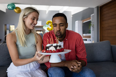 Celebrating birthday, man blowing candles on cake while woman smiling on sofa. Celebration, joy, happiness, party, friendship, festiveの写真素材