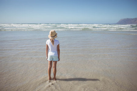 Girl standing in wet sand at shoreline wearing straw sun hat and gazing at ocean waves. Coast, seaside, leisure, relaxation, natural, outdoor, lifestyleの写真素材