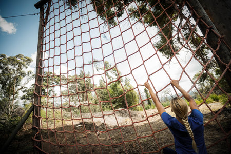 Woman gripping red rope net on wooden posts, preparing to climb wooded obstacle course, copy space. Adventure, fitness, outdoor, challenge, determination, strength, staminaの写真素材
