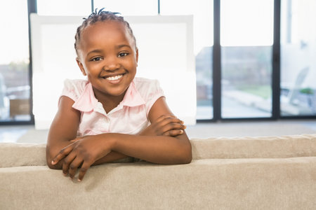 African American girl leaning over sofa back smiling at camera in living room through window panels. Child, interior, natural light, brightness, home, cheerful, casualの写真素材