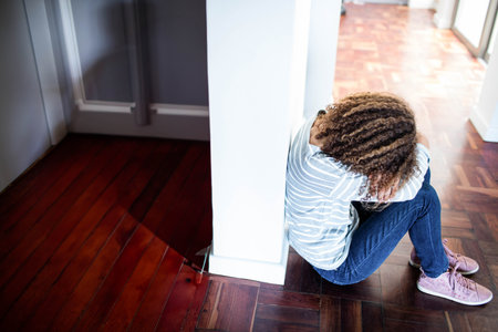 Teenage girl curling knees, resting head on arms beside white column on parquet floor in corridor. Youth, solitude, introspection, interior, minimalist, comfort, emotionalの写真素材