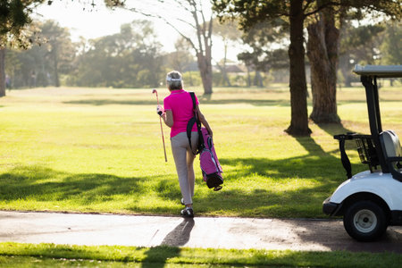 Mature female golfer walking along fairway path in pink polo shirt carrying bag and holding club. Athletic, leisure, outdoor, sport, elegance, serenity, lifestyleの写真素材