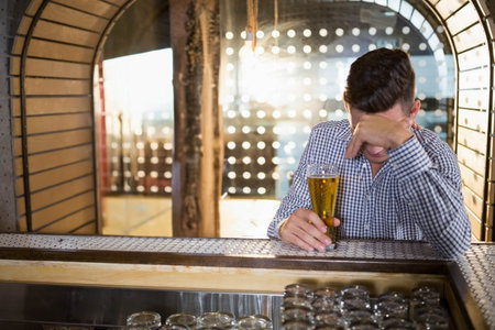 Man wearing checkered shirt leaning on metal mesh bar counter holding pint glass, copy space. Contemplative, moody, rustic, introspection, evening, solitary, ambianceの写真素材