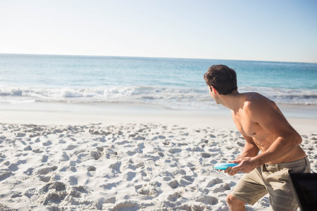 Shirtless male standing on beach by turquoise sea, preparing to throw light blue disc, copy space. Beach, recreation, leisure, outdoor, sport, relaxation, vitalityの写真素材