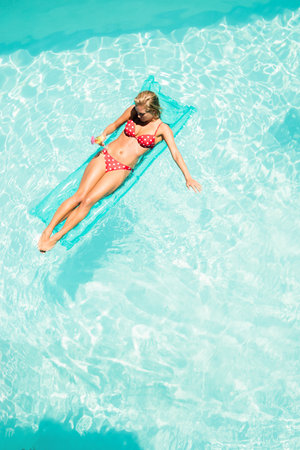 Female reclining on aqua inflatable mattress in residential pool holding beverage glass, copy space....