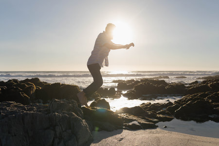 Man jumping barefoot between jagged rocks on beach at low tide wearing striped shirt and trousers. Adventure, exploration, dynamic, nature, minimalist, active, ruggedの写真素材