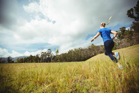 Woman wearing blue tee and dark leggings running up meadow slope under cloudy sky, copy space. Athletic, outdoor, scenic, fitness, movement, nature, healthyの写真素材