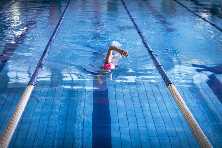 Senior female swimmer gliding through tiled pool lane by lane ropes in pink swim cap goggles. Athlete, fitness, aquatic, training, water sport, outdoor, vibrantの写真素材