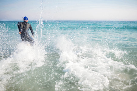 Male athlete striding into turquoise sea wearing neoprene wetsuit and blue swim cap, copy space. Adventure, water sports, ocean, athleticism, dynamic, outdoor, active lifestyleの写真素材