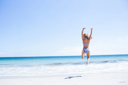Female beachgoer jumping mid-air on sandy beach under clear blue sky with turquoise ocean waves. Active, leisure, outdoor, vibrant, adventure, fitness, summerの写真素材