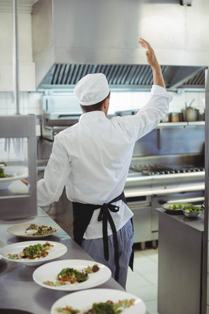 Mid adult male chef raising right hand and inspecting dishes on steel counter in restaurant kitchen....
