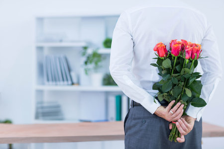 Mature man standing in office holding pink-orange rose bouquet behind back near desk, copy space. Professional, corporate, workspace, elegant, motivational, refined, floralの写真素材