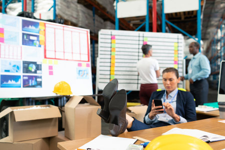 Female manager sitting on desk using smartphone among boxes, boards in warehouse, copy space. Logistics, teamwork, planning, industrial, collaboration, organization, efficiencyの写真素材