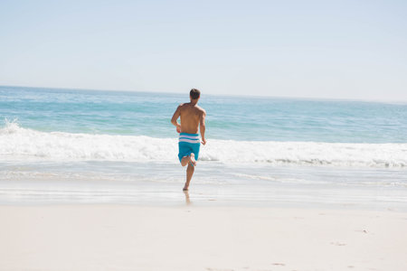 Shirtless male running toward waves on sandy shoreline wearing blue and white striped swim trunks. Athletic, leisure, outdoor, nature, movement, recreation, landscapeの写真素材