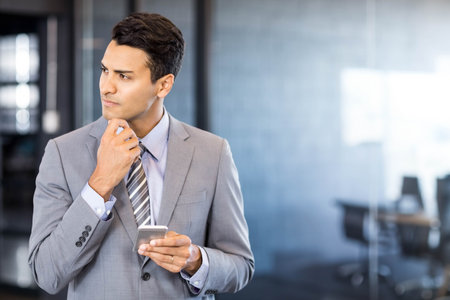 Business professional holding smartphone and touching chin thoughtfully in office with glass walls. Corporate, modern, professional, workspace, technology, strategy, consultingの写真素材