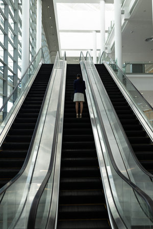 Asian woman riding up escalator in modern atrium holding handbag by glass railings. Modern, corporate, architecture, professional, elegance, interior, urbanの写真素材