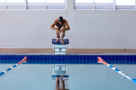 Male swimmer crouching on starting block labeled 9 at pool, wearing black swim cap and goggles. Athleticism, water sports, competition, training, aquatic, body fitness, sportswearの写真素材