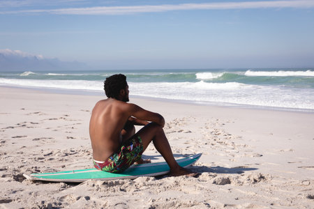 African American male sitting cross legged on turquoise surfboard on white sand gazing at waves. Adventure, leisure, vibrant, serenity, purposeful, outdoor, explorationの写真素材