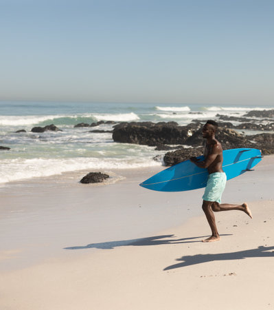 African American man sprinting on sandy shore with waves carrying bright blue surfboard, copy space....