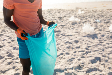 African American man collecting debris using blue trash bag on beach wearing gloves, copy space....