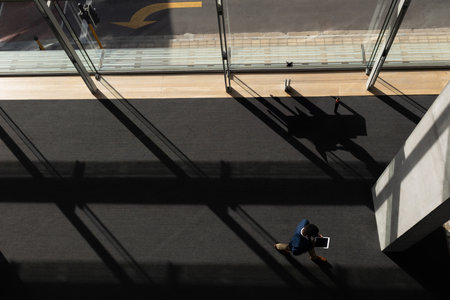 Man walking through carpeted hall toward glass doors under sunlight holding tablet, copy space. Professional, modern, architecture, urban, technology, businessの写真素材