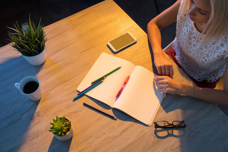Female student writing in blank notebook at study desk with pencils, smartphone and mug, copy space. Creative, workspace, feminine, illumination, organization, aesthetics, productivityの写真素材