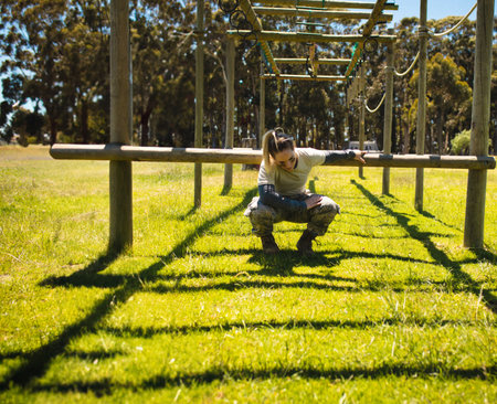Woman navigating horizontal beam obstacle on wooden posts on grassy field wearing military uniform. Adventure, fitness, strength, resilience, outdoor, training, athleticの写真素材