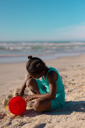 African American girl shaping sand structures on sandy shoreline using red bucket under clear sky. Child, recreation, playful, serenity, outdoor, vibrant, leisureの写真素材