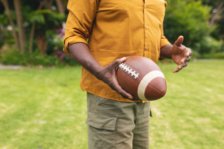 Senior man wearing mustard shirt holding brown football in garden. Resilience, vitality, outdoor, leisure, healthy lifestyle, nature, casualwearの写真素材