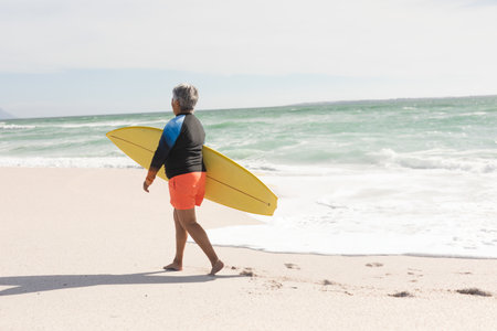Senior man walking barefoot on sandy beach wearing blue rash guard and carrying yellow surfboard. Coastline, adventure, outdoor, vitality, serenity, exploration, landscapeの写真素材