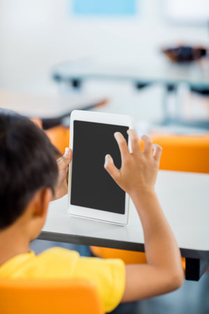 White tablet being held upright on white desk with blank screen in classroom, with orange chairs. Technology, education, innovation, learning, youth, classroom, digitalの写真素材