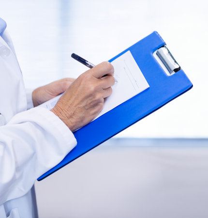 Medical worker wearing white coat with stethoscope holding clipboard filling form with pen, blinds. Medical, healthcare, professional, clinical, documentation, examination, patient careの写真素材