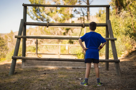 Boy standing with hands on hips facing large wooden log obstacle frame in sunlit forest clearing. Adventure, exploration, playground, outdoor, sport, activity, youthfulの写真素材
