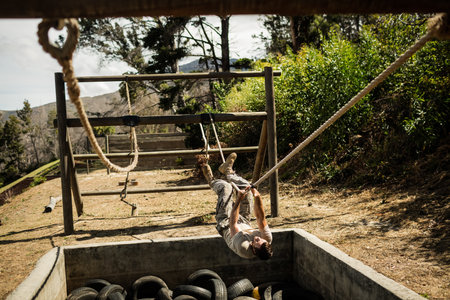 Male trainee swinging on rope across tire pit on outdoor obstacle course under bright sky. Adventure, endurance, outdoortraining, rugged, athleticism, resilience, natureの写真素材