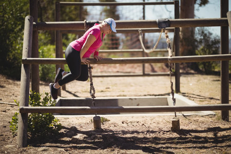 Athlete vaulting over wooden three-beam obstacle in sunlit forest clearing with hanging chains. Outdoor, fitness, agility, adventure, natural, activewear, sportの写真素材