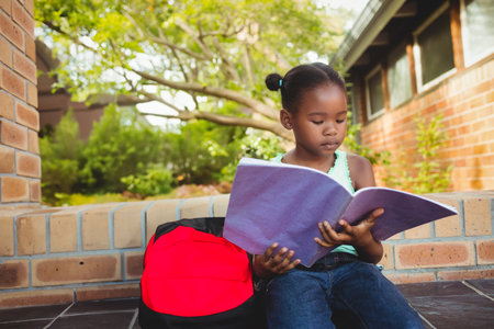 Child reading large purple book on low brick ledge outside building, with red and black backpack. Outdoor, educational, vibrant, youthful, literature, relaxationの写真素材