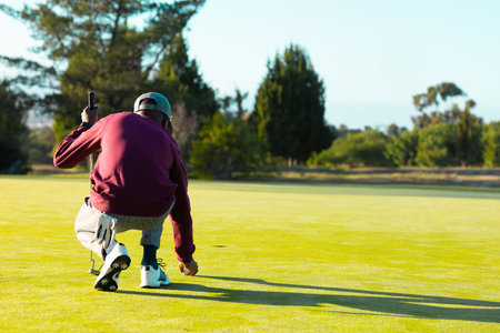 African American man with cap crouching on putting green placing golf ball with putter, copy space....