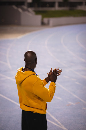 African American man wearing yellow hoodie standing on track edge at stadium, copy space. Athletics, sport, activewear, outdoors, motivation, endurance, competitiveの写真素材
