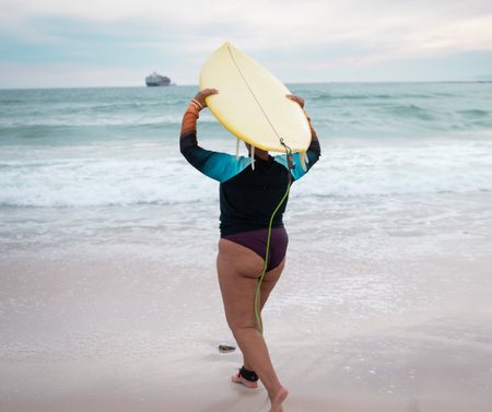 African American woman walking into surf carrying yellow surfboard wearing rash guard with leash. Ocean, adventure, outdoors, activity, dynamic, nature, coastalの写真素材