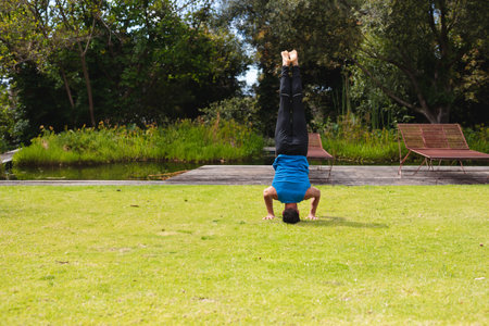 Man wearing sportswear performing headstand on grass in private garden near wooden deck and pond....