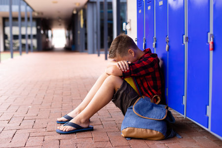 School-aged boy sitting on school corridor leaning against blue lockers with backpack, copy space. Student, youth, adolescence, outdoor, casual, education, urbanの写真素材