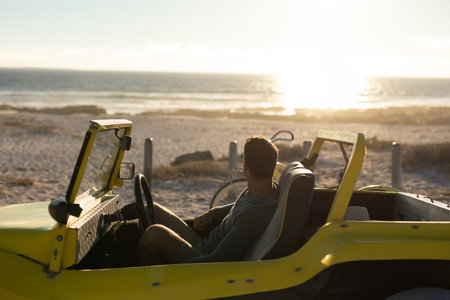 Man sitting in yellow dune buggy on sandy beach, gazing toward ocean horizon with bicycle. Adventure, leisure, escape, vacation, outdoor, scenic, tranquilityの写真素材