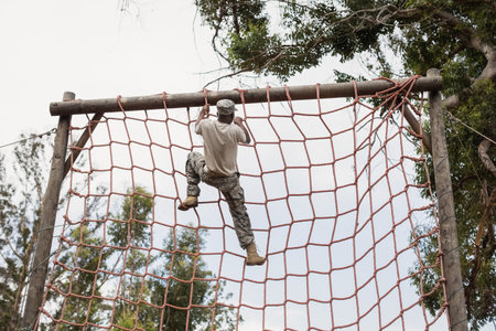 African American male soldier in camo climbing red rope net reaching wooden beam on obstacle course. Adventure, fitness, resilience, outdoor, athletic, training, challengeの写真素材