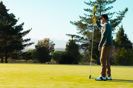 Gloved male golfer standing on putting green holding flagstick, focusing on ball at cup, copy space. Athletic, leisure, outdoor, scenic, concentration, landscape, vibrantの写真素材