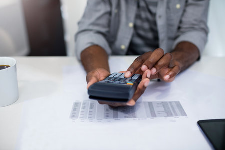 African American man holding calculator, reviewing spreadsheet at office with coffee mug and tablet. Business professional, financial analysis, data management, modern workspace, productivity, corporate, technologyの写真素材