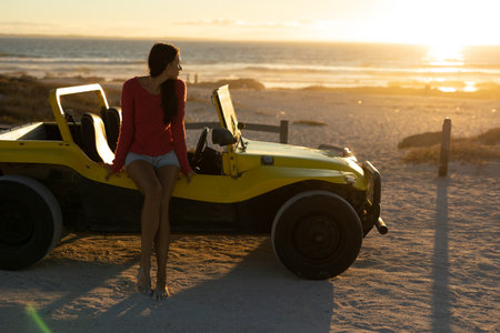 Woman sitting on yellow dune buggy at beach at sunset, gazing ocean and posts, copy space. Vacation, adventure, leisure, relaxation, scenic, outdoor, vibrantの写真素材